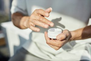 Close up on hands of mature man holding plastic jar of beauty cream applying nourishing product on skin while taking care of face in bathroom, copy space